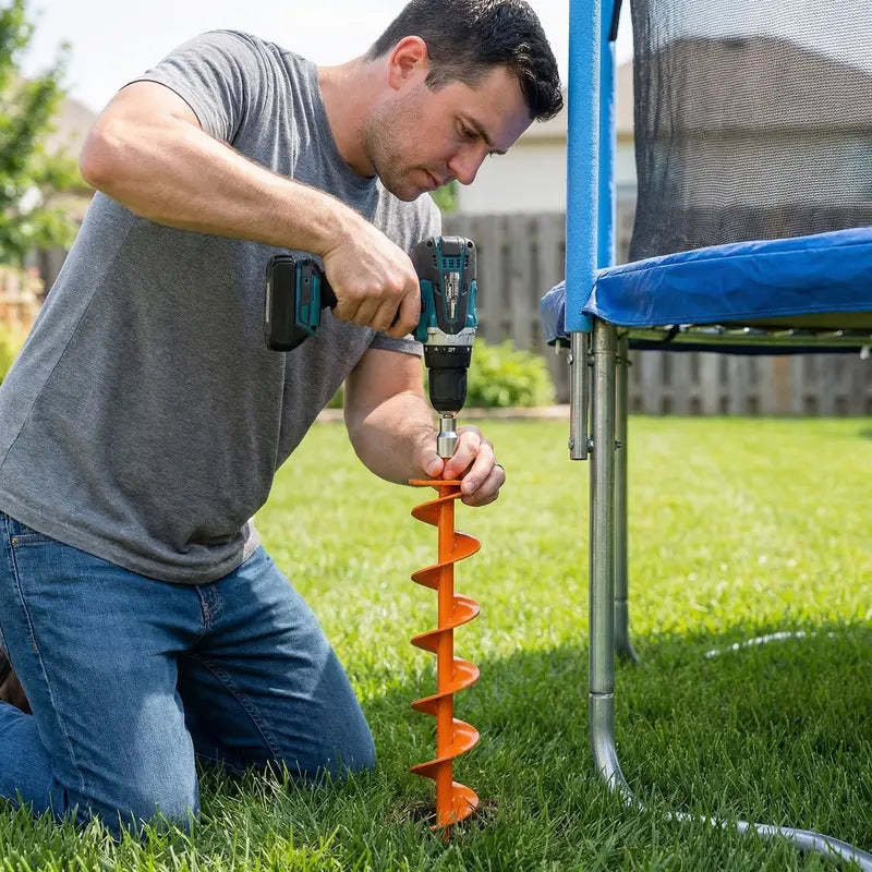 Person using a cordless drill to install an orange full-spiral ground anchor beside an outdoor trampoline